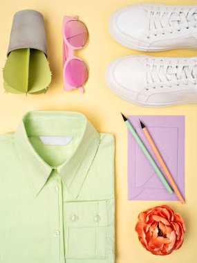 Top view of mint green plain cotton formal shirt, lavender envelopes, white sneakers and artificial paper succulent on a pastel yellow background. Feminine work outfit or minimalist office concept.