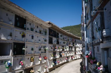 Riomaggiore, İtalya - Nisan 2022: İtalyan mezarlığında beyaz hücreli ve her bir hücrenin yanında çiçekler bulunan Kolombiya. Cinque Terre Ulusal Parkı 'ndaki mezarlıkta iki sıra mezar hücresi arasında geçiş.
