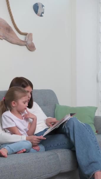 A young mother reads a book on the couch for her little blonde daughter — Stock Video