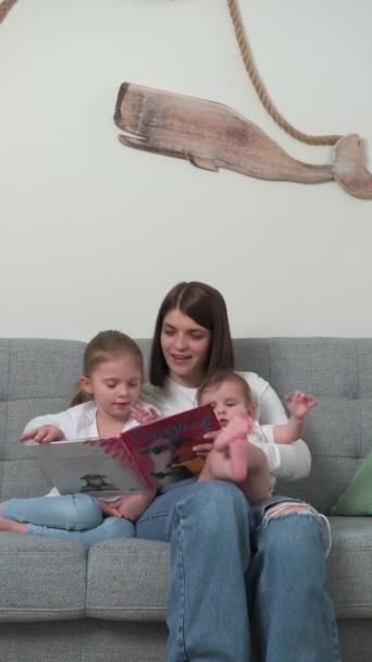 Una joven madre con un hijo y una hija pequeños lee un libro sentado en el sofá — Vídeo de stock