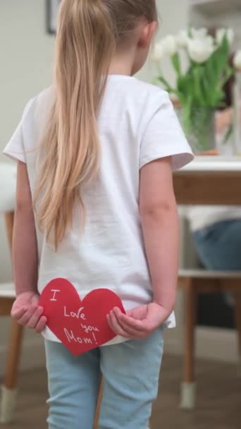 En liten blond flicka håller ett kort i form av ett hjärta för sin mamma bakom ryggen — Stockvideo