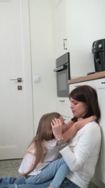 Sarışın bir kızı olan bir anne mutfak zemininde oturup konuşur.