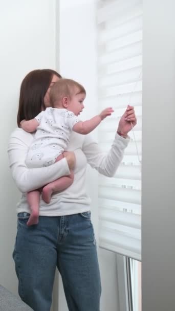 Een jonge moeder met een zoontje in haar armen staat bij een raam in een heldere kamer — Stockvideo