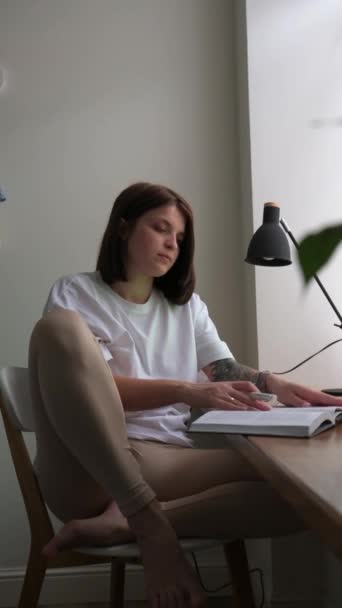Una joven se sienta con un libro junto a la ventana y trabaja o estudia a distancia — Vídeos de Stock