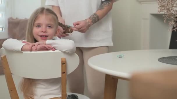 妈妈为坐在白色椅子上的金发小女儿编辫子 — 图库视频影像