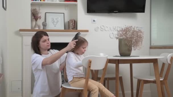 Η μαμά πλέκει μαλλιά για την μικρή ξανθιά κόρη που κάθεται σε μια λευκή καρέκλα. — Αρχείο Βίντεο
