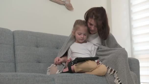 En mamma med en liten dotter sitter på soffan och leker på tabletten insvept i en filt — Stockvideo