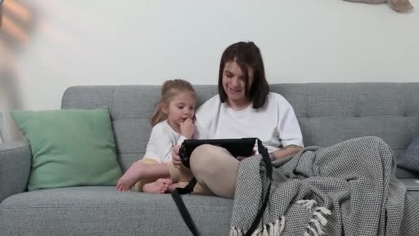 Uma mãe com uma filha pequena senta-se no sofá e brinca no tablet embrulhado em um cobertor — Vídeo de Stock