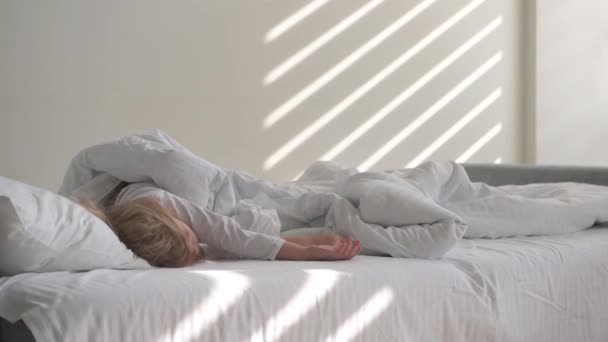 Una niña rubia de 4 años duerme en una cama blanca en los rayos del sol en la pared — Vídeos de Stock