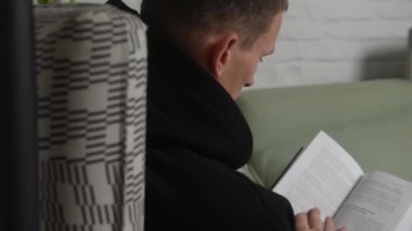 a young man in black clothes flips through a book sitting on a sofa in an interior with white walls. High quality FullHD footage
