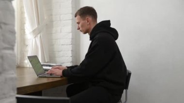 Siyah eşofmanlı genç bir adam dizüstü bilgisayar ve kitapla çalışıyor.