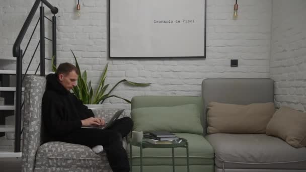 En ung kille sitter nära trappan i lägenheten och skriver något på sin bärbara dator — Stockvideo