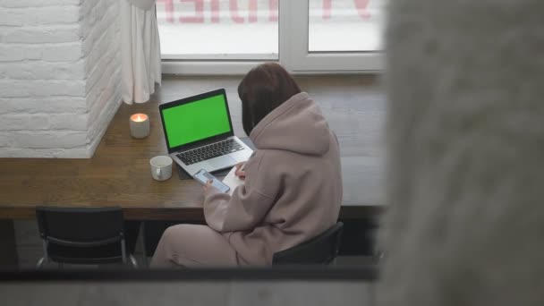 Top view of a girl who sits near a large window and works on a laptop with a green screen — Vídeo de Stock