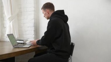 Siyah eşofmanlı genç bir adam dizüstü bilgisayar ve kitapla çalışıyor.