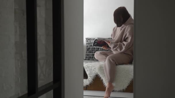 A peeped angle in which a young girl in a light suit sits on a blanket and reads a magazine in a stylish light interior in the Scandinavian style — Wideo stockowe