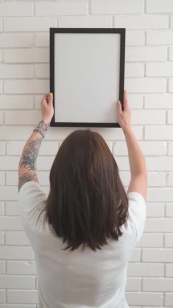 Uma menina pendura uma moldura vazia em uma parede de tijolo branco — Vídeo de Stock