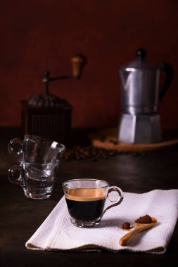 Ön planda cam bir fincanda sıcak kahve, ve çiğ kamış şekerli ahşap kaşık, arka planda antika bir kahve öğütücüsü ve kahve demliği..