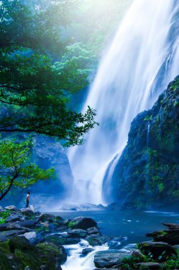 Asyalı yalınayak bir adam kayanın üzerinde duruyor ve yağmur mevsiminde büyük şelaleye bakıyor. Khlong Lan Ulusal Parkı, Tayland. Tatiller, rahatlama kavramları. Uzun pozlama.