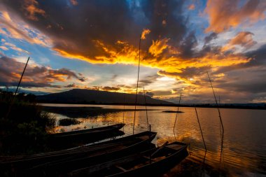 Alacakaranlıkta bir göl manzarası, dramatik bulutlar ve günbatımı gökyüzü bir göl üzerinde ve arka planda dağlar, ön planda ahşap botları olan yerel bir rıhtım. Loei, Tayland. Bir göle odaklan..