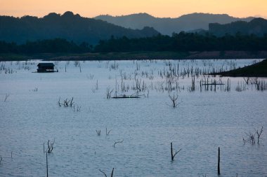 Alacakaranlıkta sakin bir göl manzarası, yüzen bir ev ve gölde geleneksel bir sal dalışı, dağlar ve arka planda günbatımı gökyüzü. Khao Laem Gölü, Tayland.