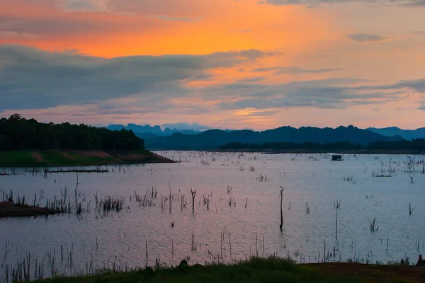 Alacakaranlıkta tropik bir göl manzarası, yüzen bir ev ve gölde geleneksel bir sal balıkçı ağı, arka planda dağlar ve günbatımı gökyüzü. Khao Laem Gölü, Tayland.