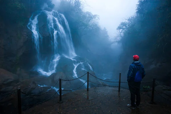 A tourist man wearing a red hat stands in front of a tropical waterfall in foggy, a tourist attraction in Sapa, Vietnam. Long exposure.