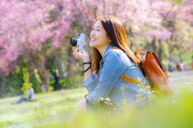 Klasik fotoğraf makinesi kullanan genç, güzel Asyalı bir gezgin. Bahar kiraz çiçeklerinin ortasında gülümseyen bir mutlu fotoğraf çek. Seyahat yaşam tarzı, dünya gezgini, Asya seyahat fikri..