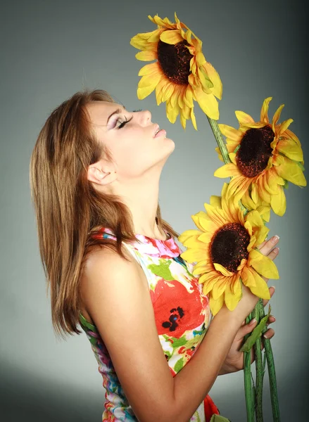 Attraktives, brünettes Mädchen in buntem Kleid, stehend mit Sonnenblumen, Studioaufnahme, auf grau — Stockfoto