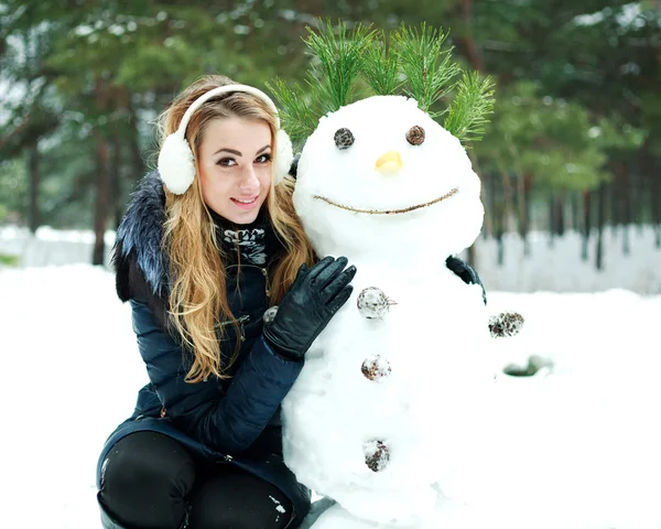 Pretty girl with snowman in pinewood on a winter day — Stock Photo, Image