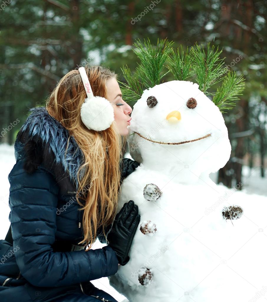 Pretty girl kissing snowman in pinewood on a winter day