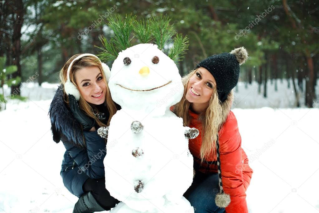 Two happy pretty girls with snowman in pinewood on a winter day