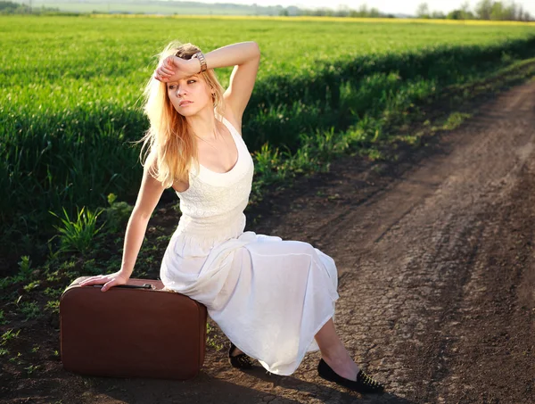 Beautiful blonde girl in long white dress, sitting on suitcase — Stock Photo, Image