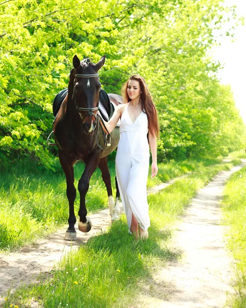 Uma menina em um vestido branco longo com um cavalo vai em uma estrada de campo — Fotografia de Stock