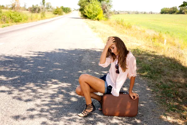 Mujer desesperada sentada en una maleta junto a un camino rural —  Fotos de Stock