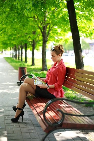 Ung kvinna sitter på bänken i grön park, läser tidningen — Stockfoto