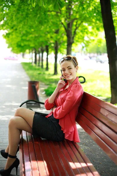 Jovem mulher sentada em um parque em um banco falando ao telefone — Fotografia de Stock