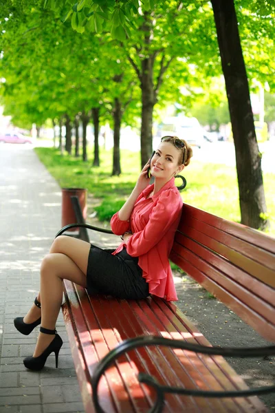 Jovem mulher sentada em um parque em um banco falando ao telefone — Fotografia de Stock