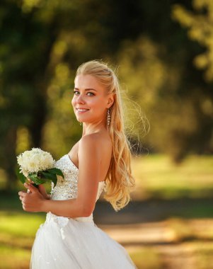 Orman parkında bir buket çiçekle uzun beyaz gelinlik içinde güzel bir gelinin portresi.