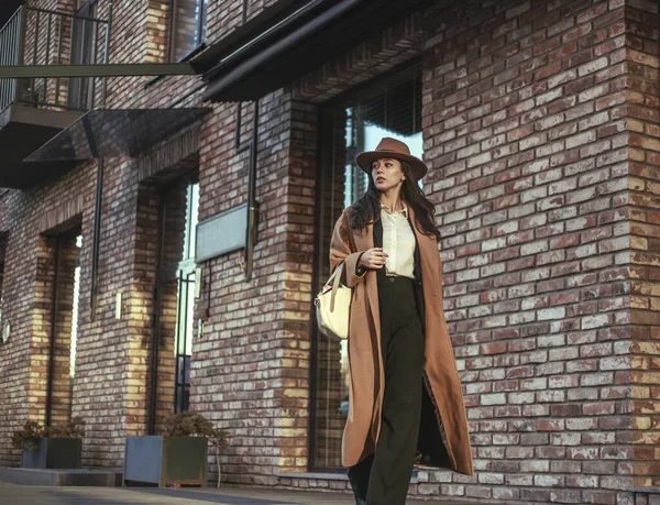 長いベージュのコートと帽子を着たアジアのビジネス女性は レンガの壁 レトロなスタイルを背景に ロフトスタイルで通りを歩いています — ストック写真