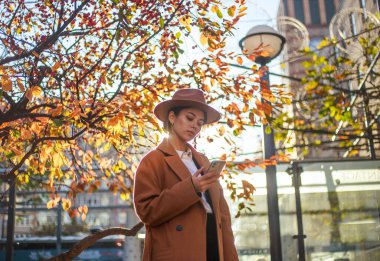 Young asian lady in beige coat and hat calls on a mobile phone on a city street in a park