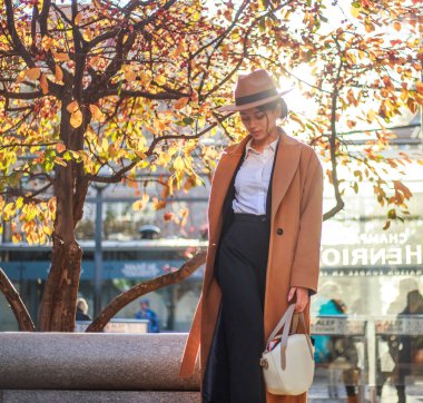 Young asian lady in beige coat and hat calls on a mobile phone on a city street in a park