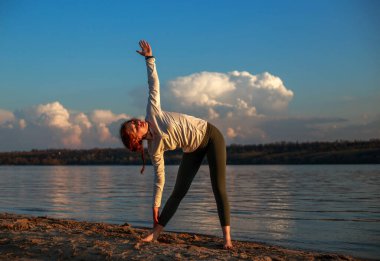 Nehrin kıyısındaki kumlu sahilde, mavi gökyüzü ve beyaz bulutların arkasında spor ve yoga yapan genç bir kadın.
