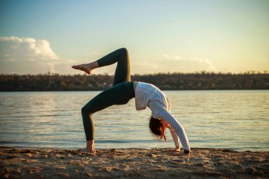 Nehrin kıyısındaki kumlu sahilde, mavi gökyüzü ve beyaz bulutların arkasında spor ve yoga yapan genç bir kadın.