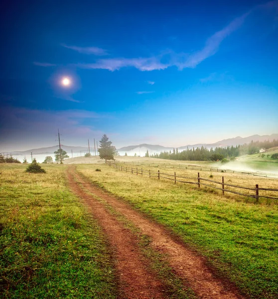 Twilight landscape, road among fields and farmland, high in the mountains, in the morning fog at dusk, on the background of the moon and blue sky