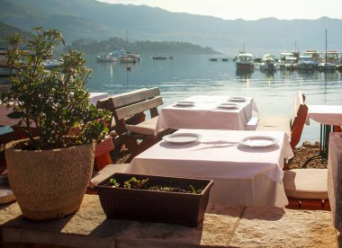 Restaurant with tables in the harbor of the coastal street overlooking the bay with yachts on a sunny summer day, Budva, Montenegro