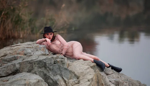 Charming young lady in a long transparent dress and a black hat in boho style sits on the shore of the lake on the stones, on the background of the reflection of reeds and forest