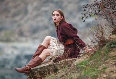 Young lady in long dress and brown boots sitting on stones, on the background of rocks and lake in autumn countryside, fashion stylish woman
