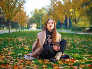 Girl in a beige coat on the background of the autumn park and yellowed leaves on the tree, thoughtfully sits on a green meadow, fashion portrait young woman
