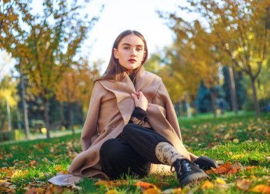 Girl in a beige coat on the background of the autumn park and yellowed leaves on the tree, thoughtfully sits on a green meadow, fashion portrait young woman