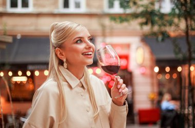 Eski bir Avrupa şehrinin akşam caddesinde bej ceketli, alımlı genç bir kadın kırmızı şarabı tadıyor ve seviyor.